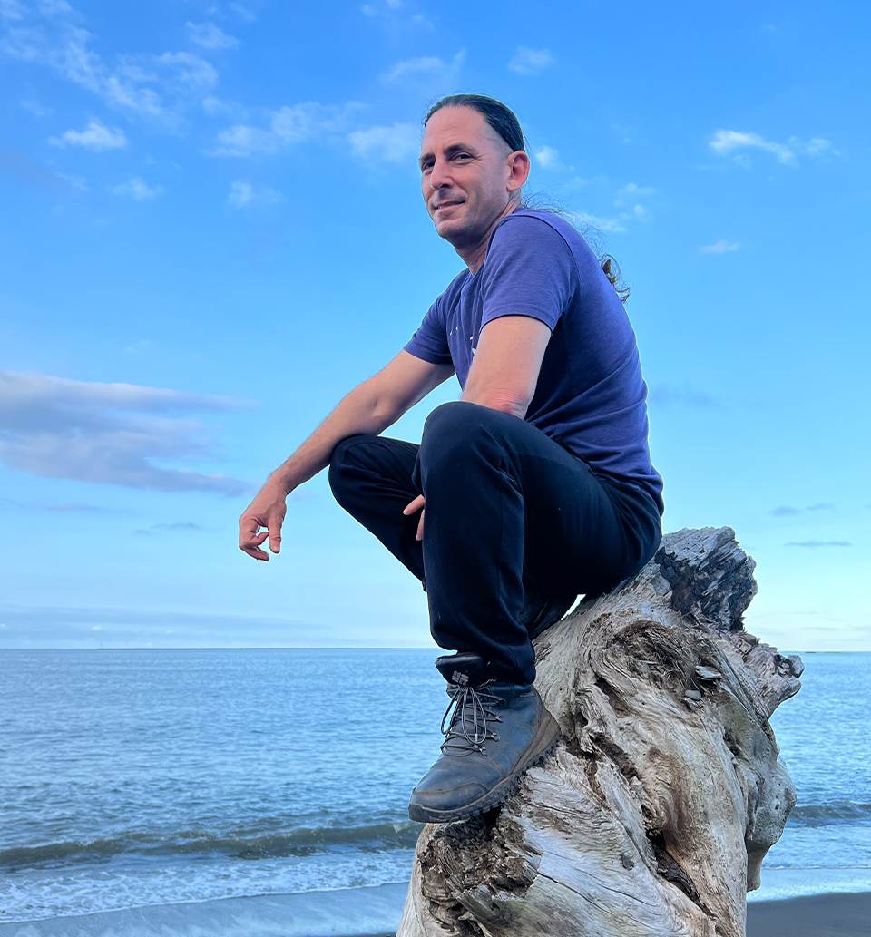 Jeff Primack in a blue shirt seated on driftwood beside the ocean